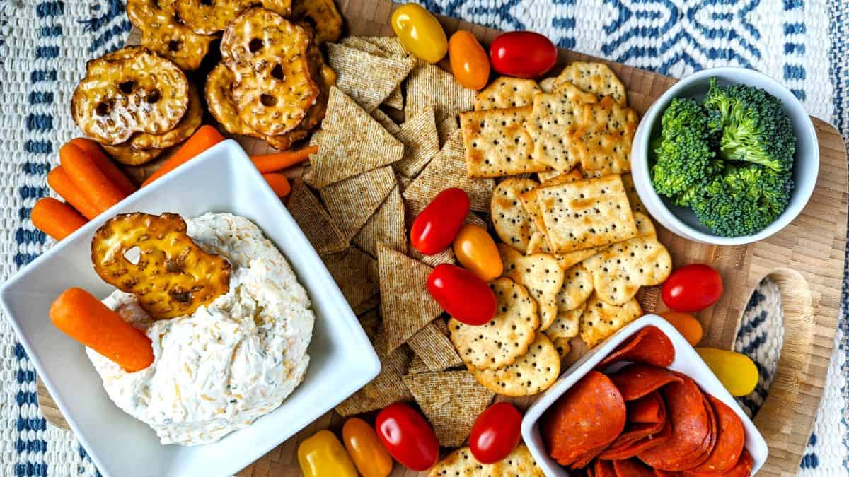A wooden board topped with crackers, pretzel crisps, sliced pepperoni, cherry tomatoes, broccoli florets, baby carrots, and a bowl of dip.