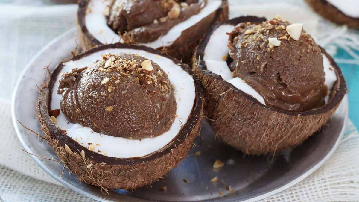 Three coconut halves filled with white cream and chocolate ice cream, garnished with chopped nuts, served on a metal plate.