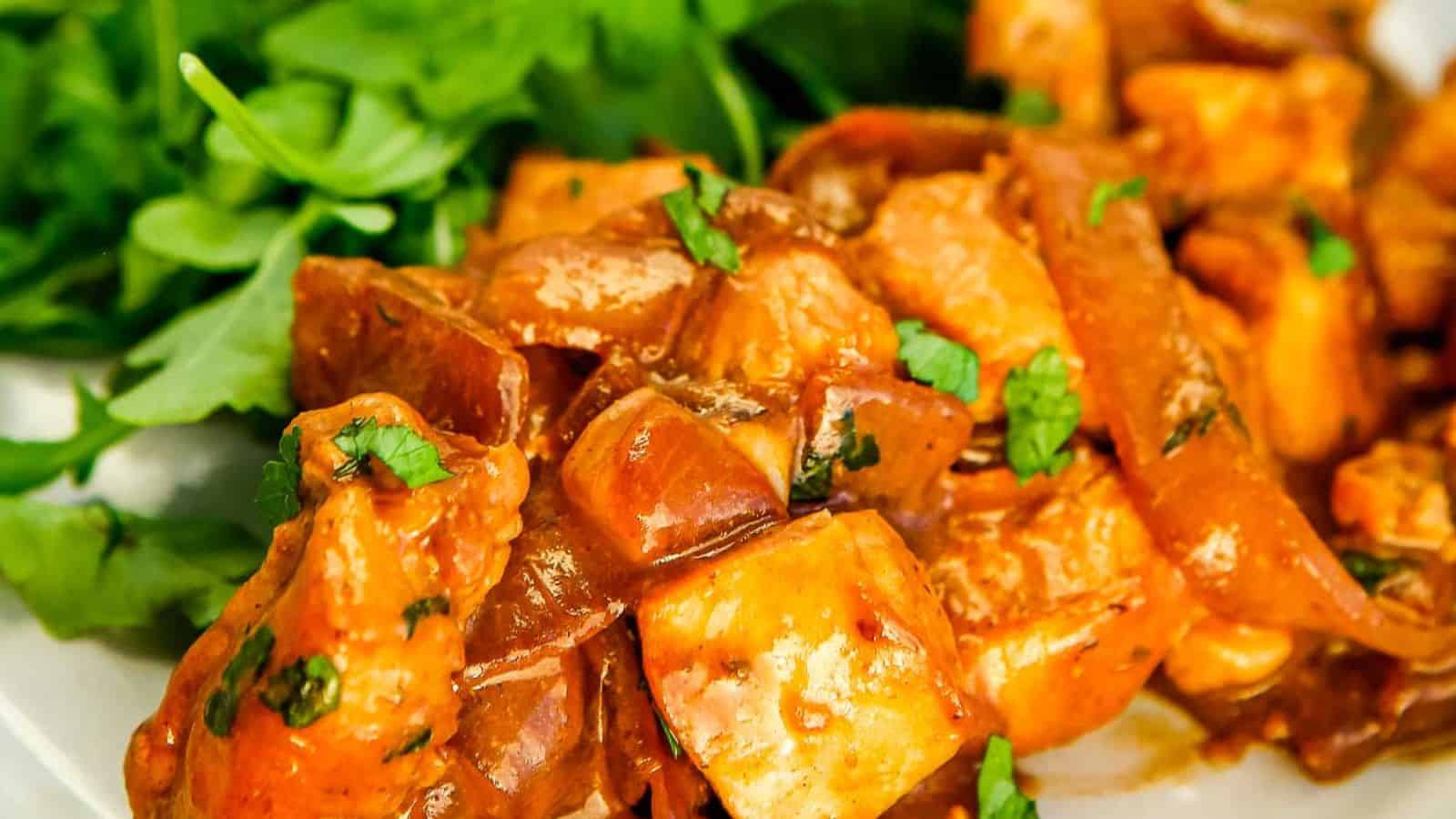 Close-up of a plate with chicken tikka masala garnished with chopped herbs, served alongside fresh green leafy vegetables.