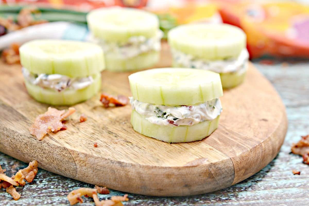 Sliced cucumber rounds filled with a creamy mixture are arranged on a wooden board, with bits of bacon scattered around.