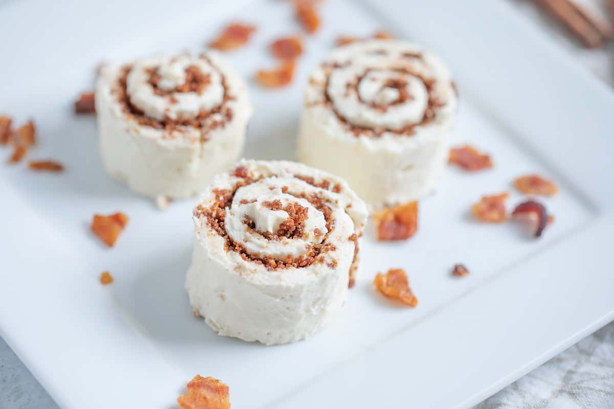 Three small cinnamon roll-shaped desserts with cream and crumb filling are arranged on a white plate, with small pieces of caramelized topping scattered around.