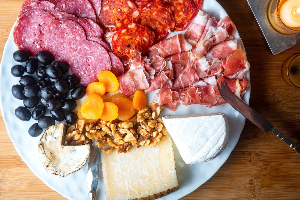 A charcuterie plate with slices of cured meats, black olives, dried apricots, walnuts, and three types of cheese on a white platter with knives on a wooden surface.