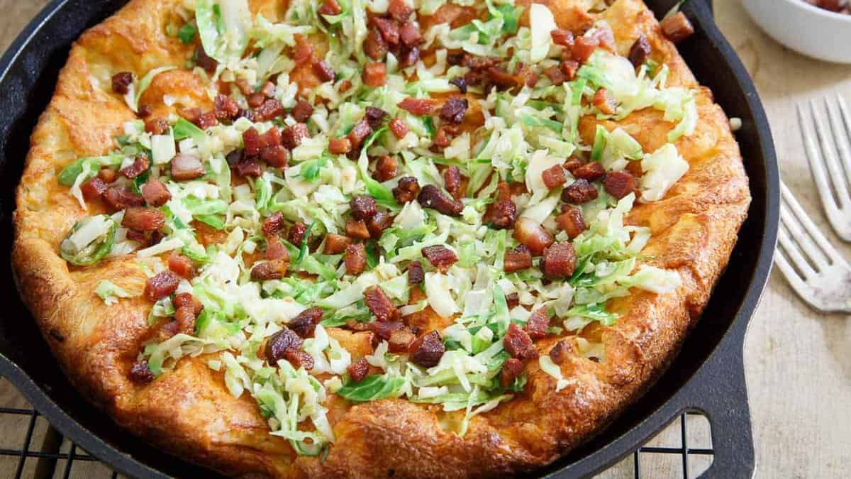 A baked pizza in a cast iron skillet topped with shredded cabbage and small pieces of cooked bacon.