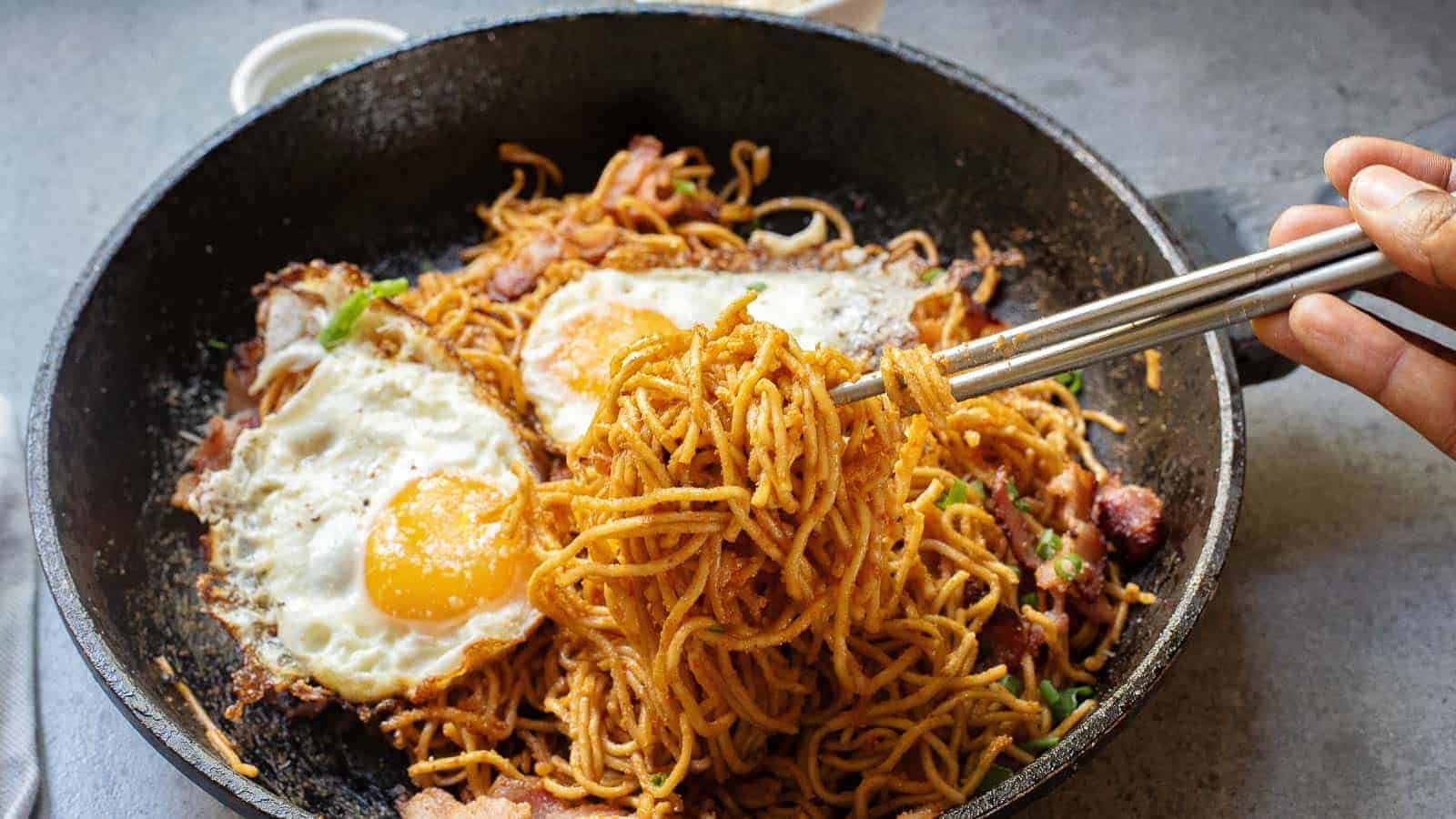 A pan of fried noodles with two sunny side up eggs, garnished with green onions, and a hand using chopsticks to lift some noodles.