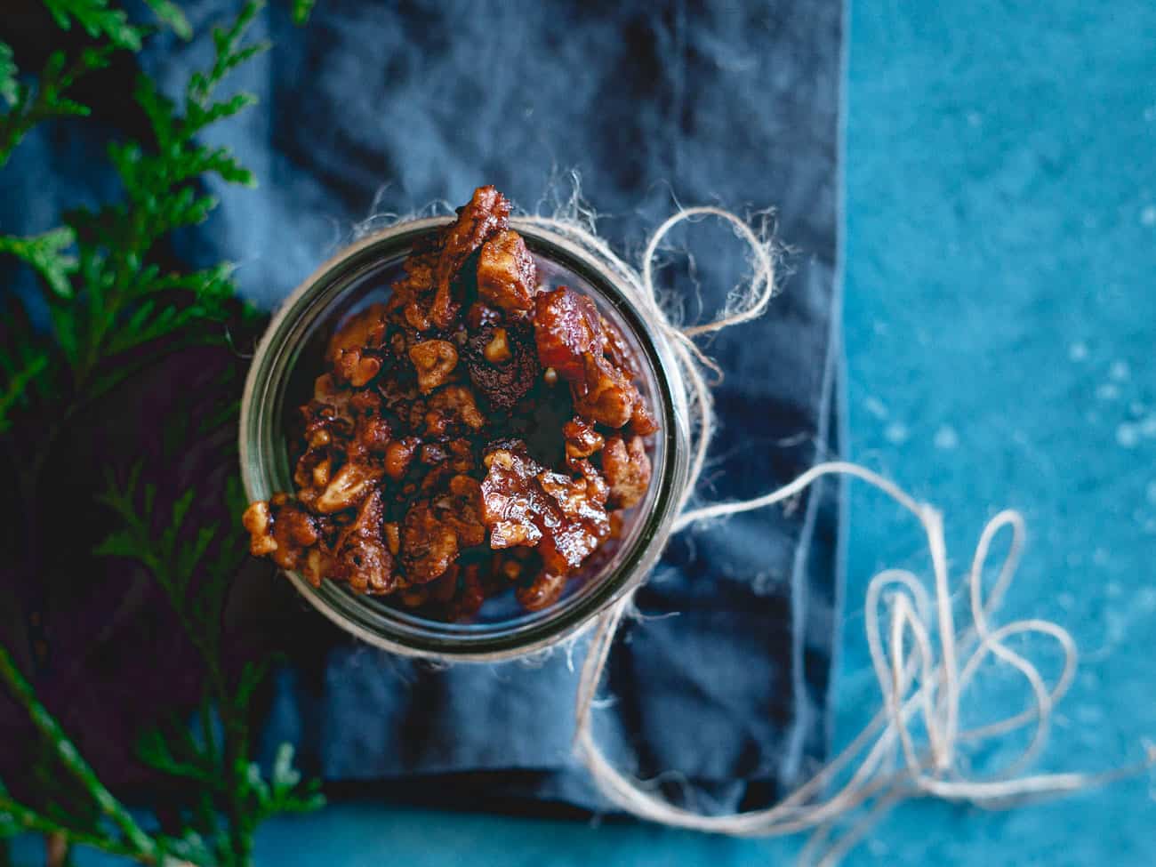 A glass jar filled with candied nuts sits on a dark cloth with twine wrapped around it, placed on a blue surface. Green foliage appears in the corner.