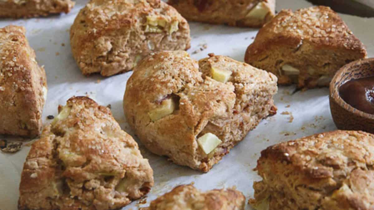 Golden brown scones with visible chunks of apple are arranged on a sheet of parchment paper, with a small wooden bowl of jam on the side.