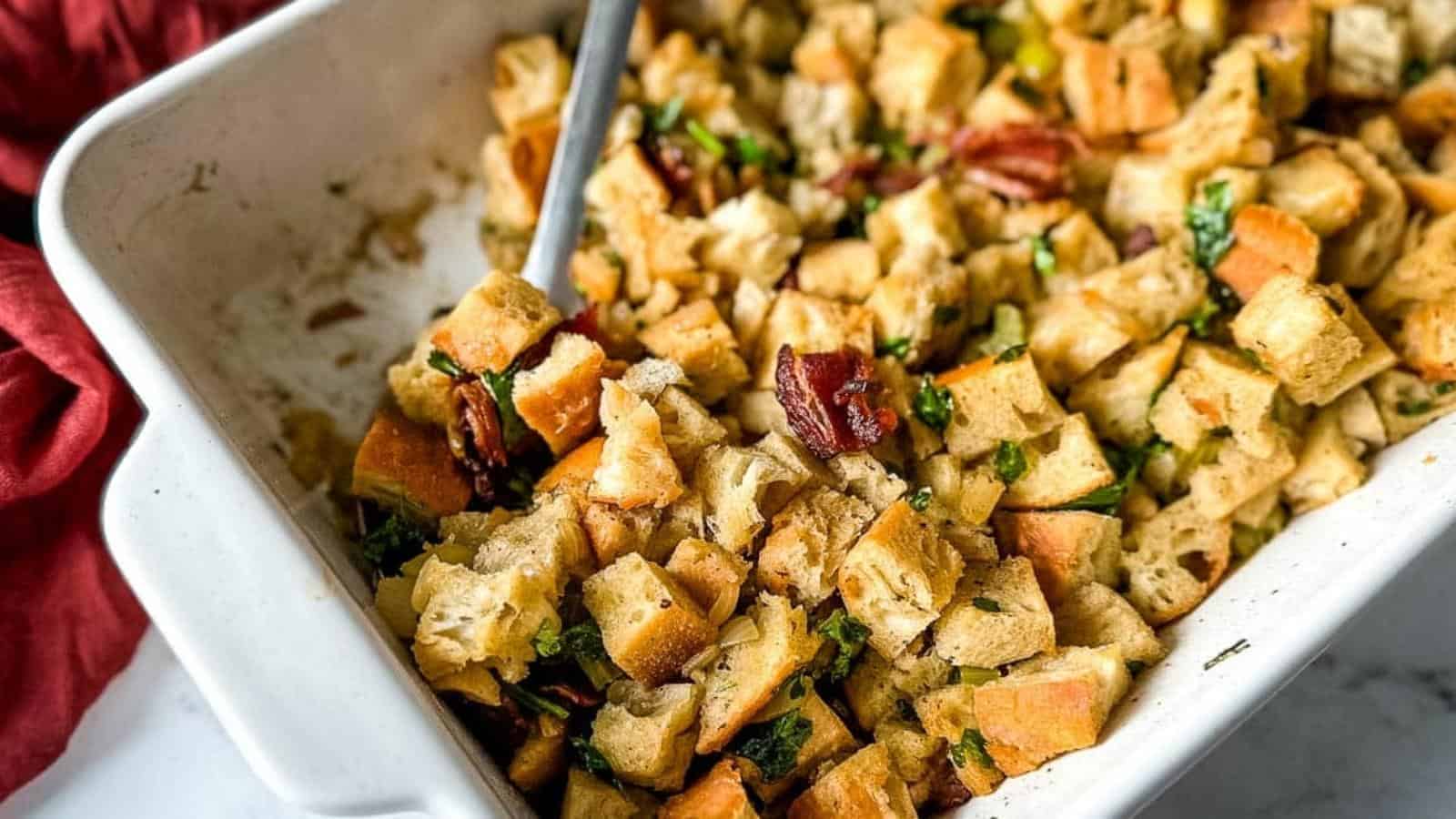 A white baking dish filled with cubed bread stuffing mixed with herbs and visible pecans, with a serving spoon inside.