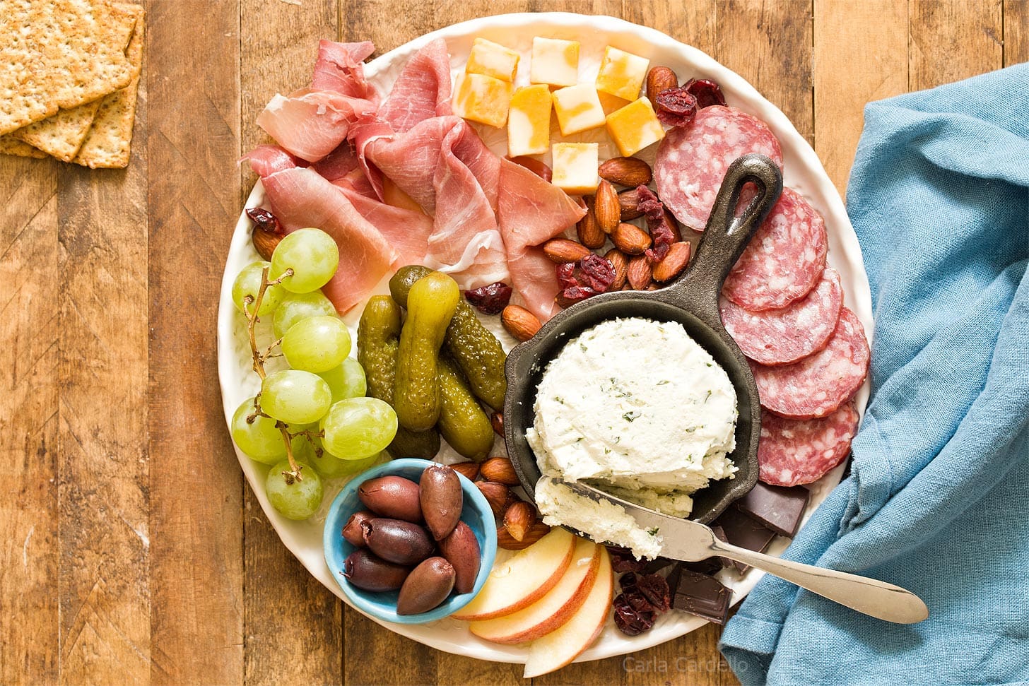 A charcuterie board with salami, prosciutto, cheese cubes, pickles, grapes, apple slices, olives, nuts, crackers, and a small skillet of soft cheese.