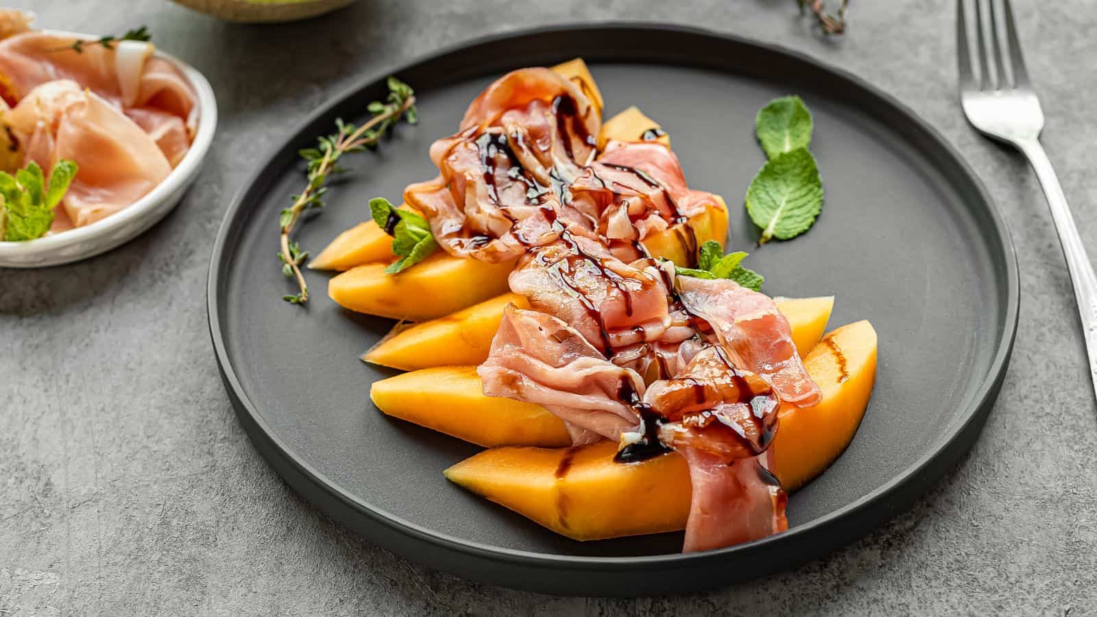 Sliced cantaloupe topped with prosciutto and balsamic glaze, garnished with fresh mint, served on a black plate.