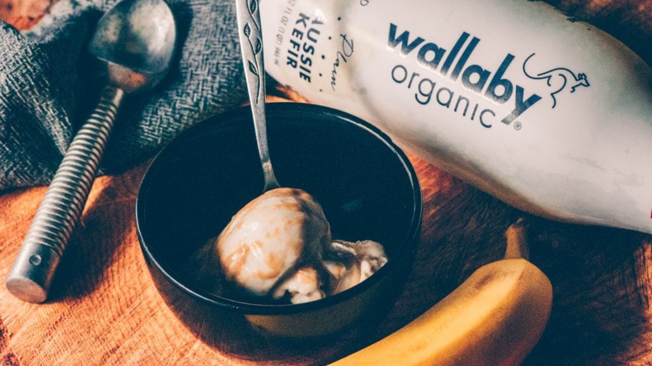 A bowl of yogurt with syrup and a spoon, a banana, and a bottle labeled Wallaby Organic on a wooden surface with a metal spoon and cloth nearby.
