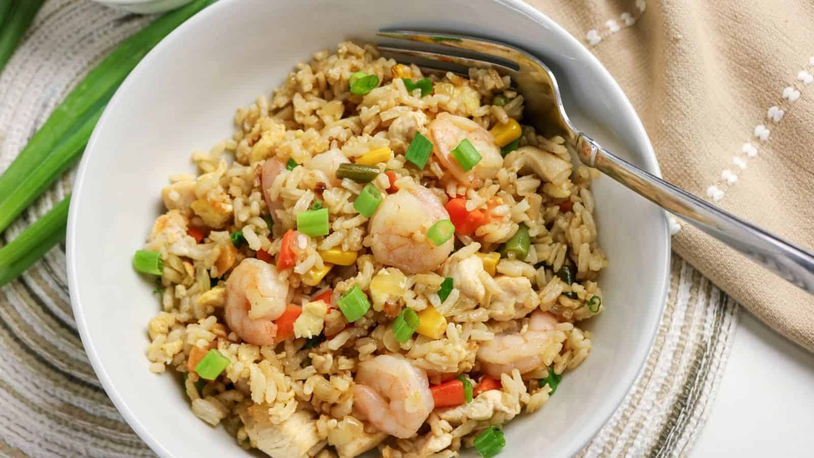 A white bowl filled with shrimp fried rice, mixed with vegetables and topped with chopped green onions, with a fork resting on the side.