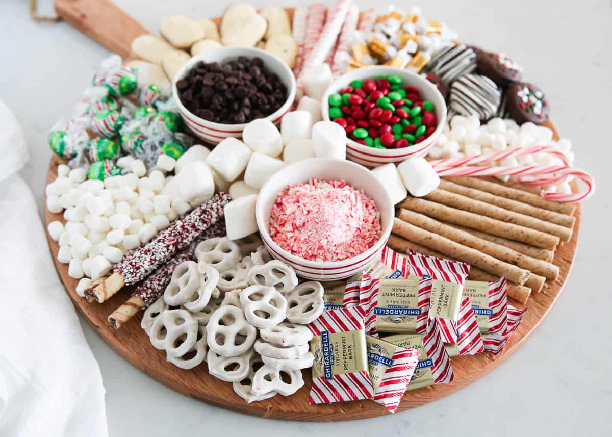 A wooden board topped with a variety of holiday sweets, including marshmallows, chocolate candies, peppermint sticks, wafer rolls, and chocolate-covered pretzels.