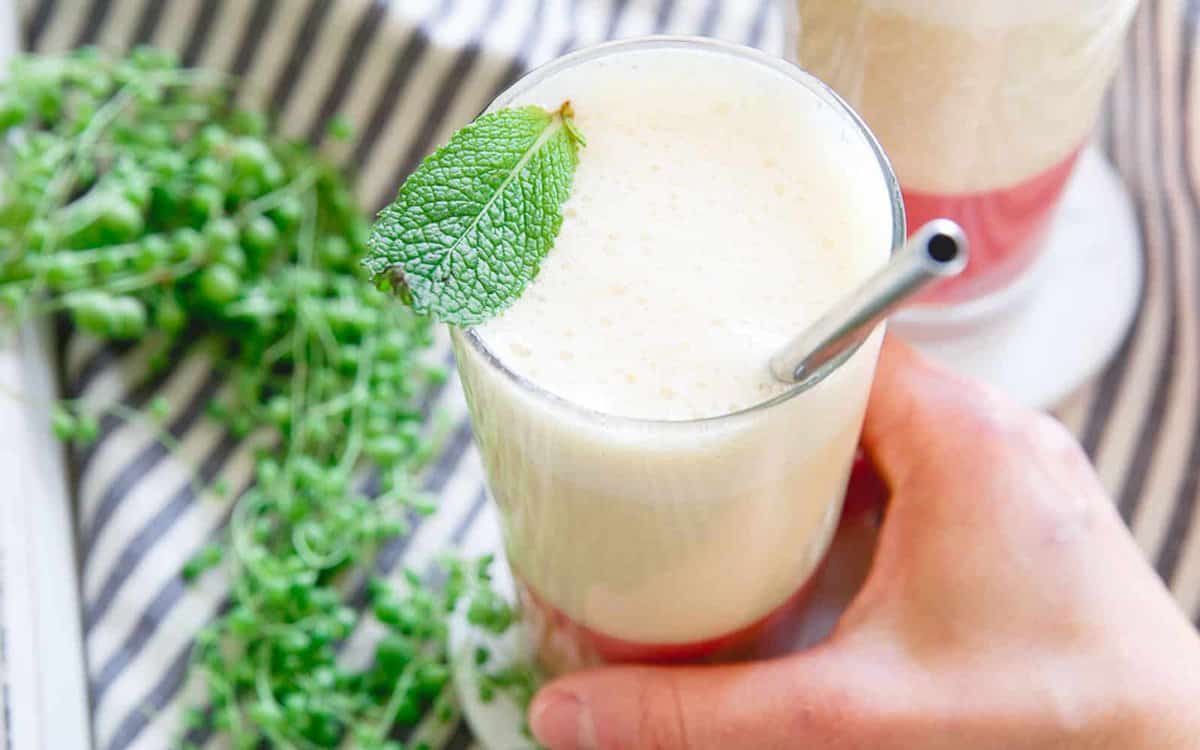 A hand holds a layered beverage in a tall glass with a metal straw and mint leaf garnish, placed on a striped surface next to green herbs.