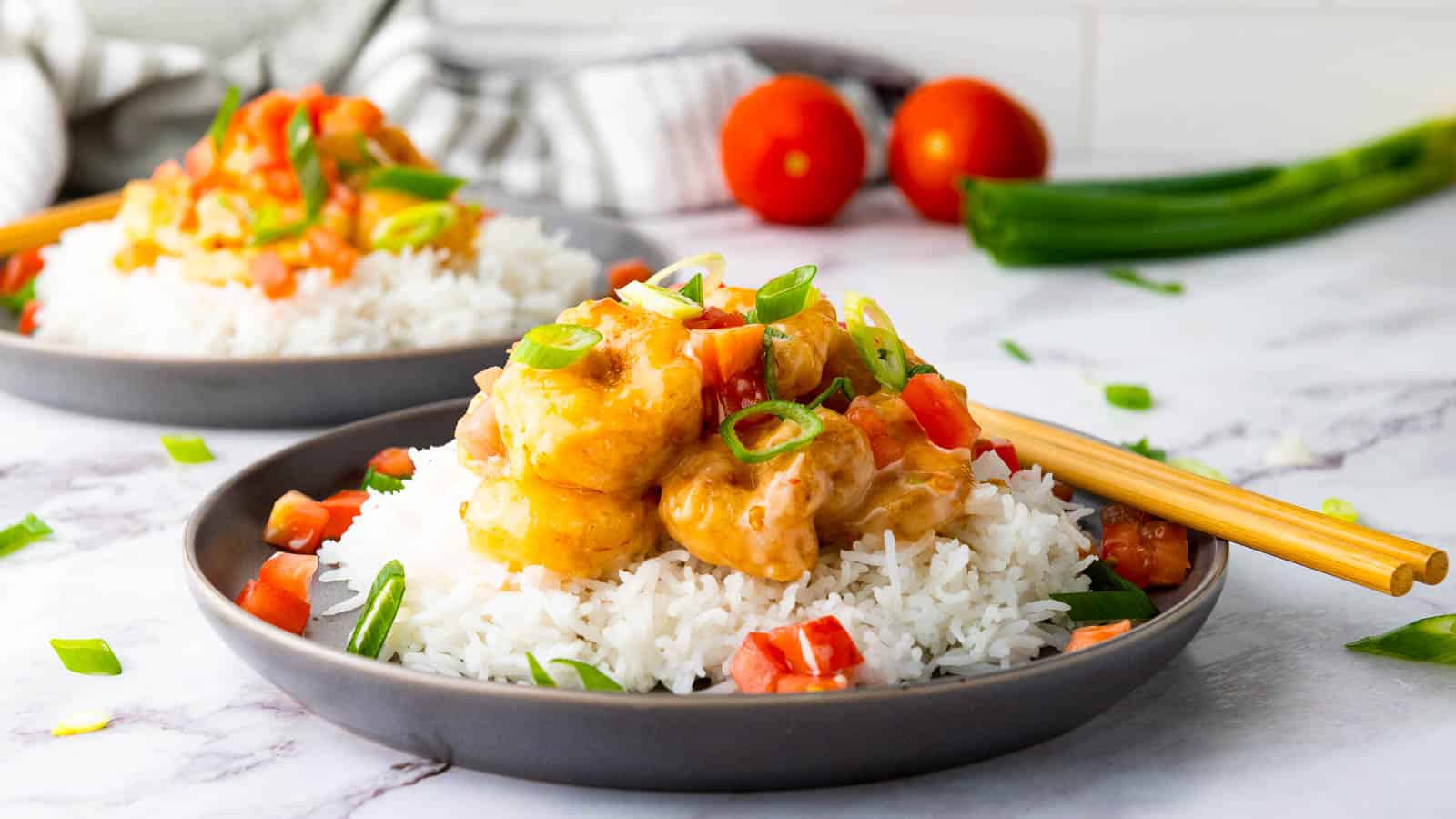 A plate of white rice topped with creamy shrimp, chopped tomatoes, and green onions, with chopsticks resting on the side. Another similar plate and vegetables are in the background.