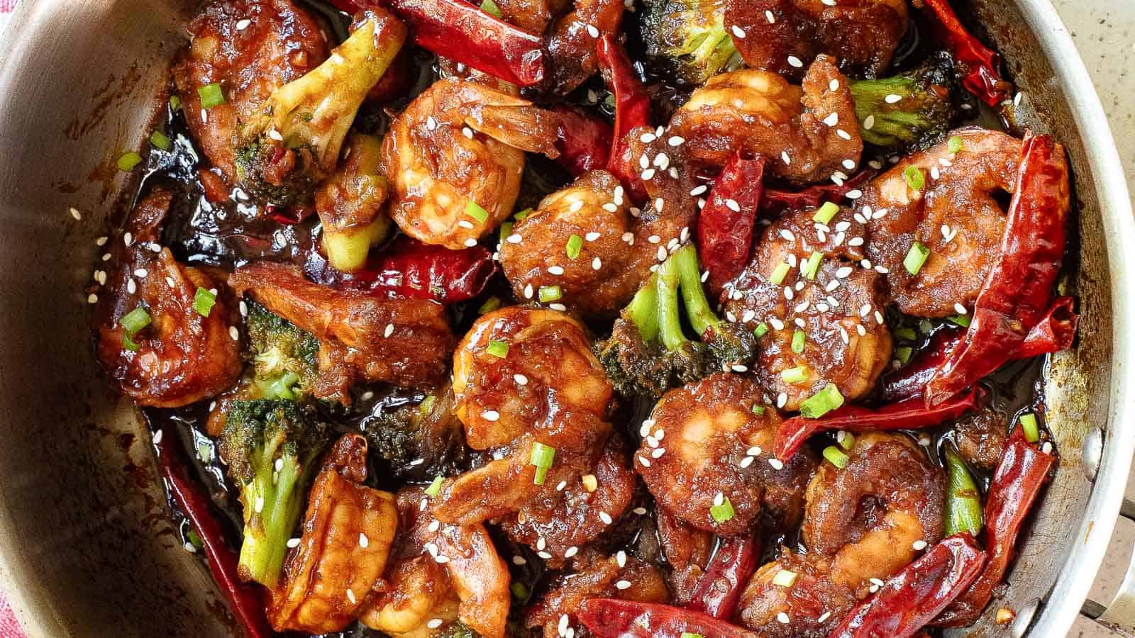 A pan filled with cooked shrimp, broccoli, dried red chili peppers, and a dark sauce, topped with sesame seeds and chopped green onions.