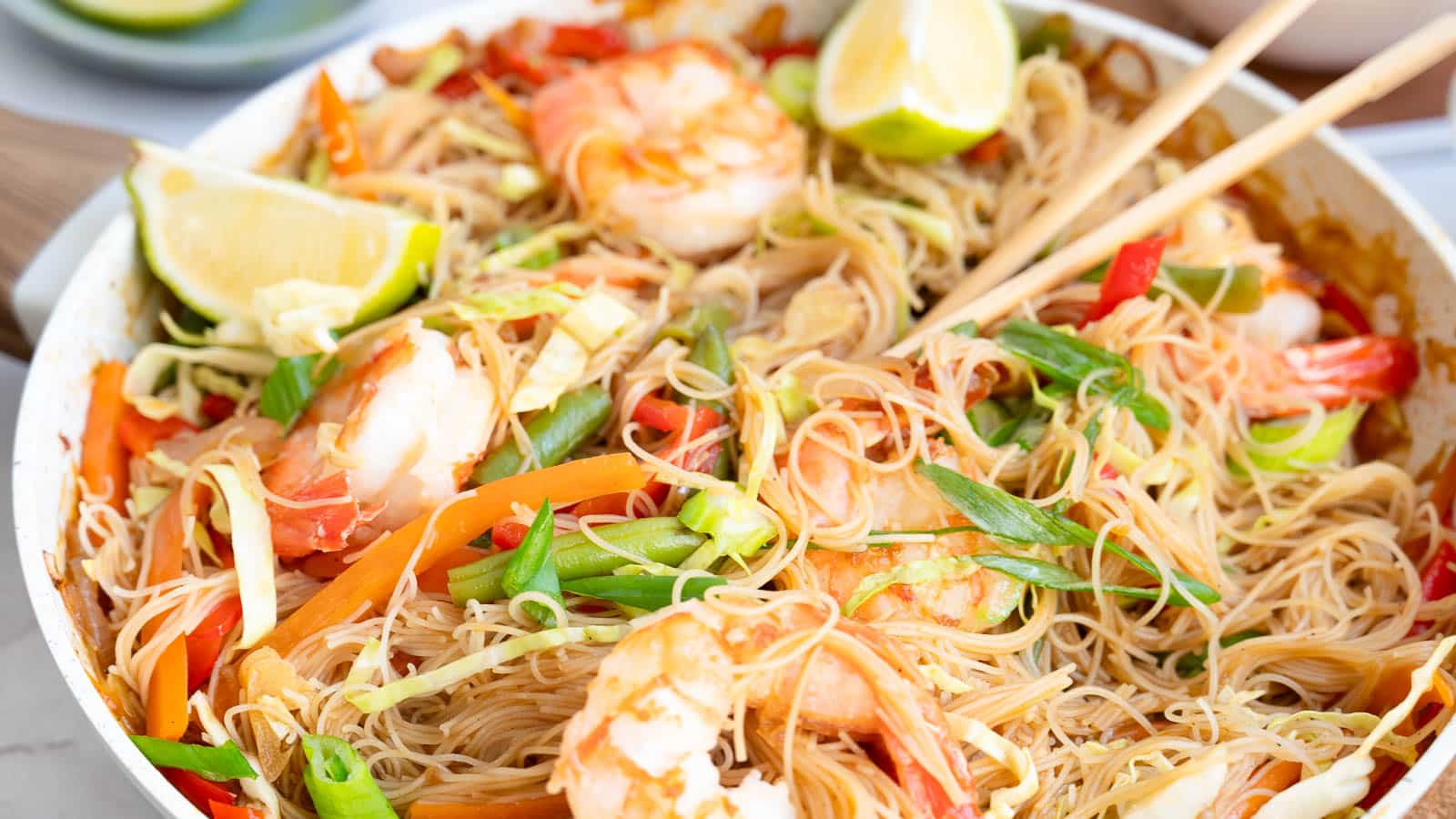 A plate of shrimp stir-fried rice noodles with vegetables, garnished with lime wedges and green onions, and a pair of chopsticks resting on top.