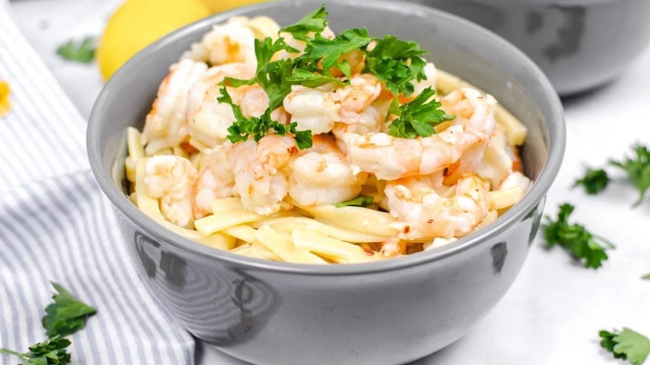 A gray bowl filled with pasta topped with cooked shrimp and garnished with fresh parsley, placed on a white surface with a striped napkin nearby.
