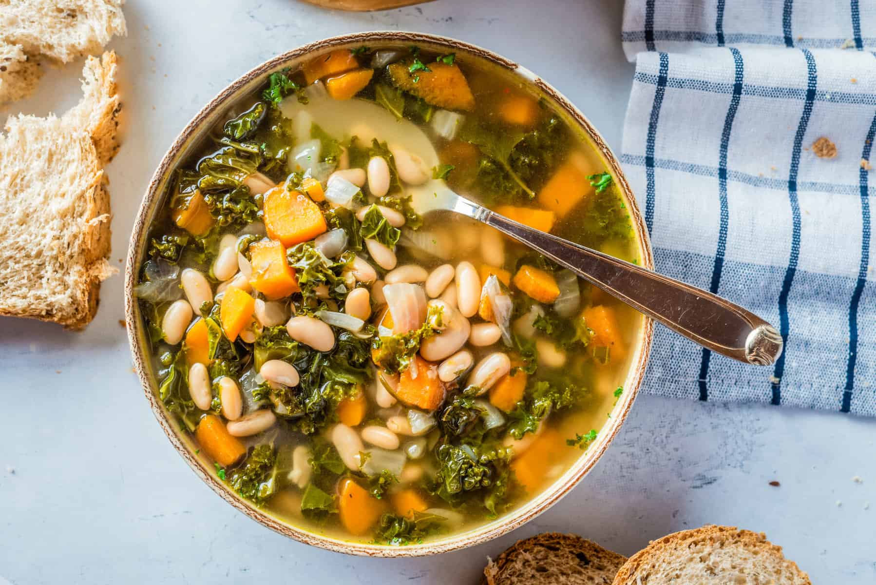 A bowl of vegetable soup with white beans, kale, and carrots sits on a table next to slices of bread and a checkered napkin, with a spoon in the bowl.