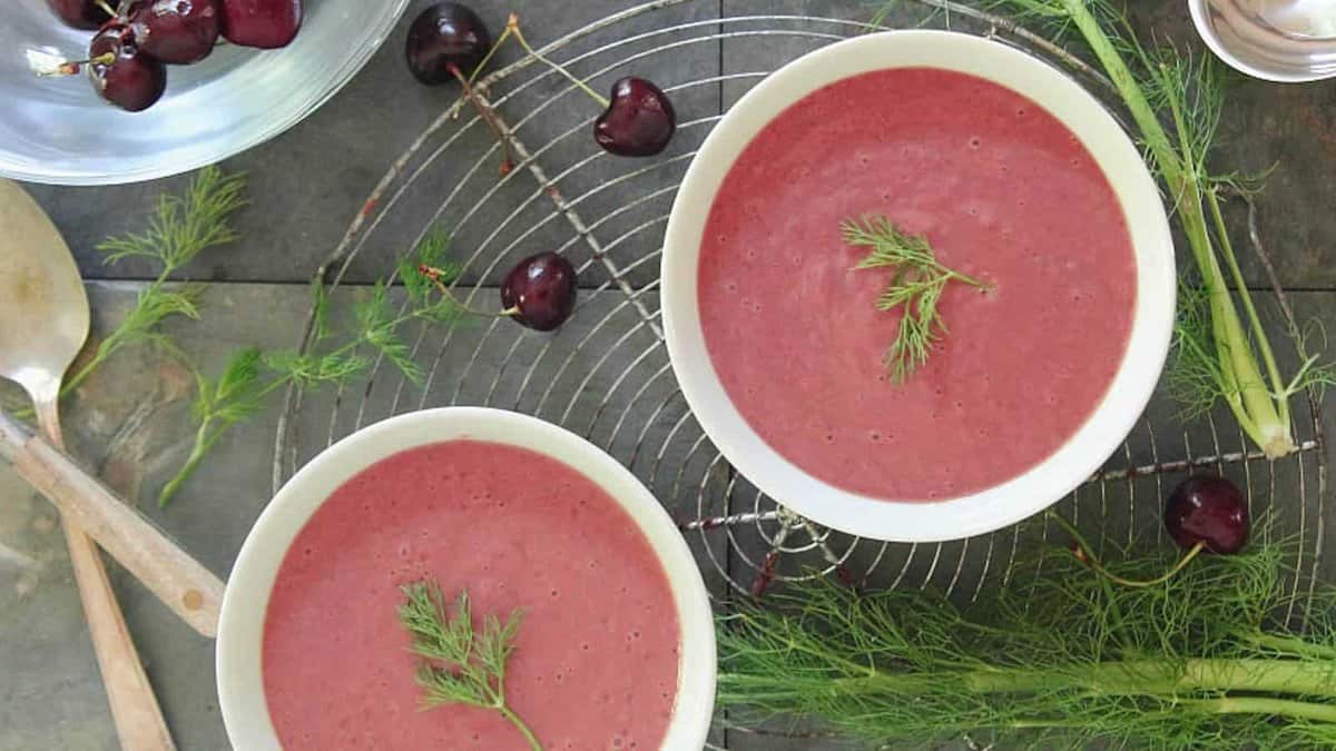 Two bowls of pink soup garnished with dill on a cooling rack, surrounded by fresh dill and whole cherries.