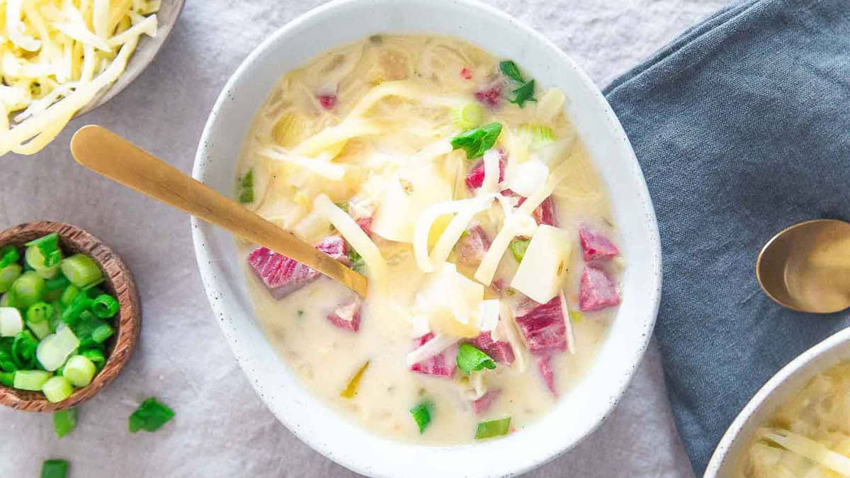 A bowl of creamy soup with chunks of corned beef, shredded cheese, and chopped green onions, placed on a light surface next to a gray napkin and a spoon.