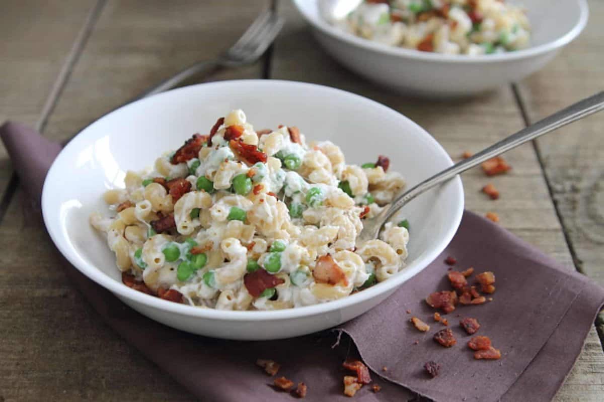 A white bowl of macaroni salad with peas, bacon, and a creamy dressing sits on a brown napkin with a fork; another bowl is in the background.