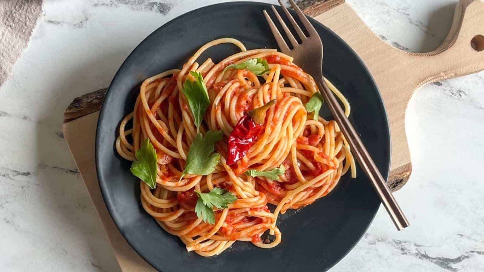A black plate of spaghetti with tomato sauce, garnished with parsley and dried chili peppers, sits on a wooden board with a fork on the side.