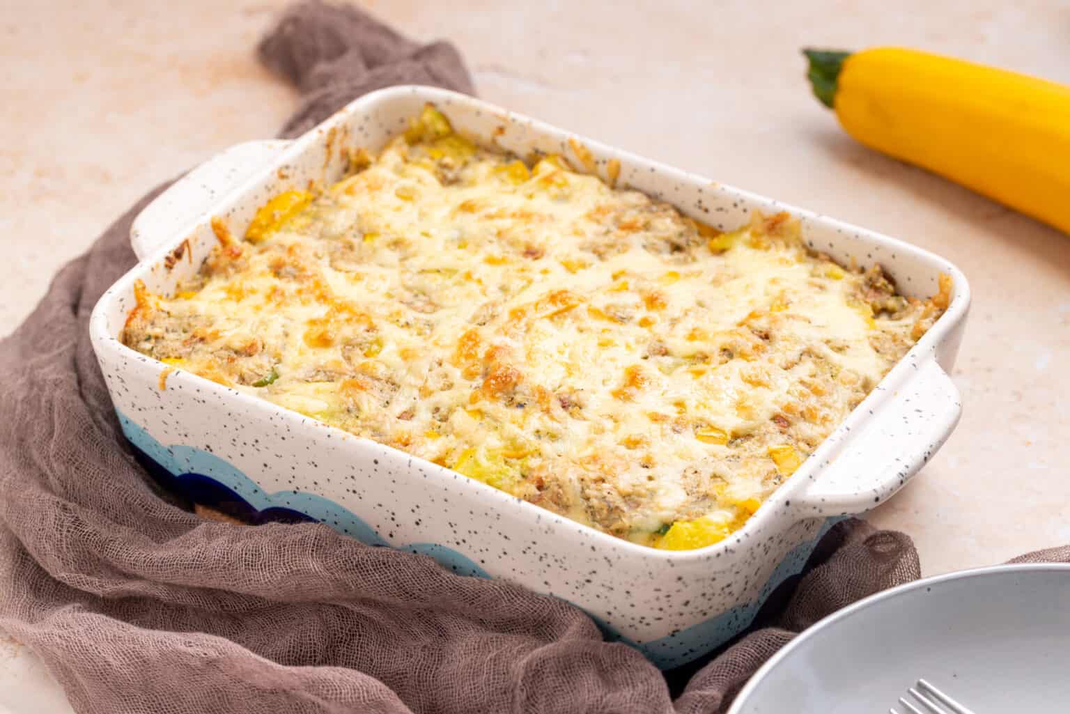 A baked casserole topped with melted cheese in a rectangular dish, with a yellow squash and a brown cloth nearby on the counter.