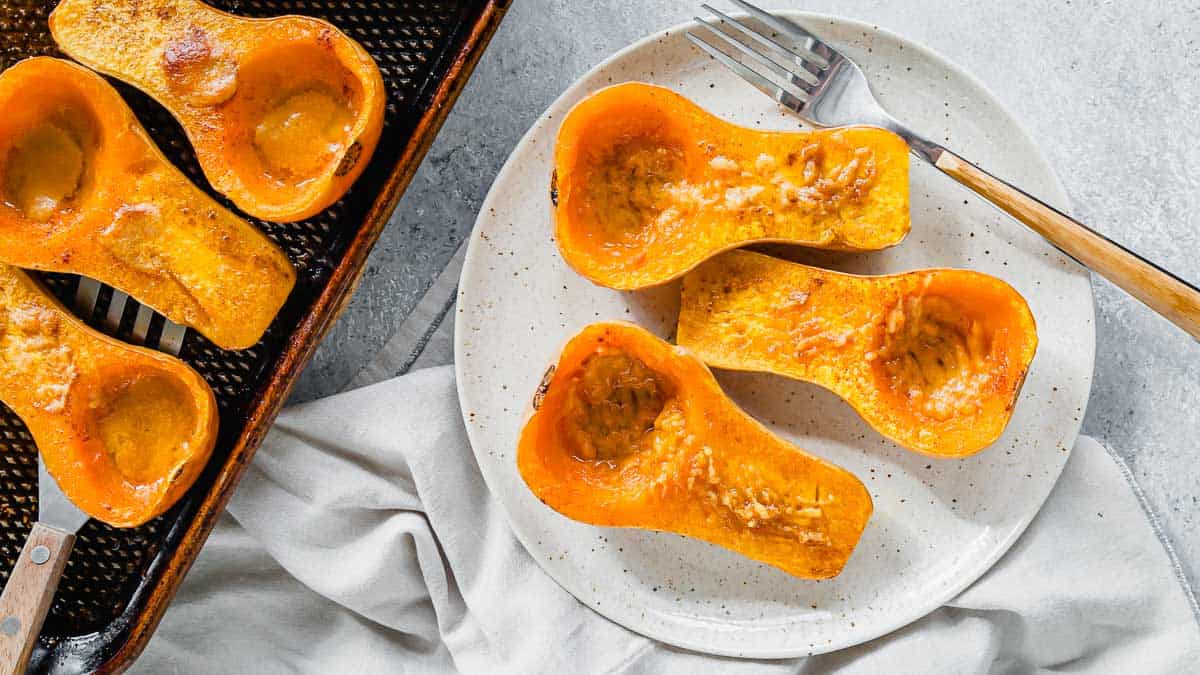 Three roasted butternut squash halves on a white plate next to a baking tray with more roasted squash halves, forks placed nearby.