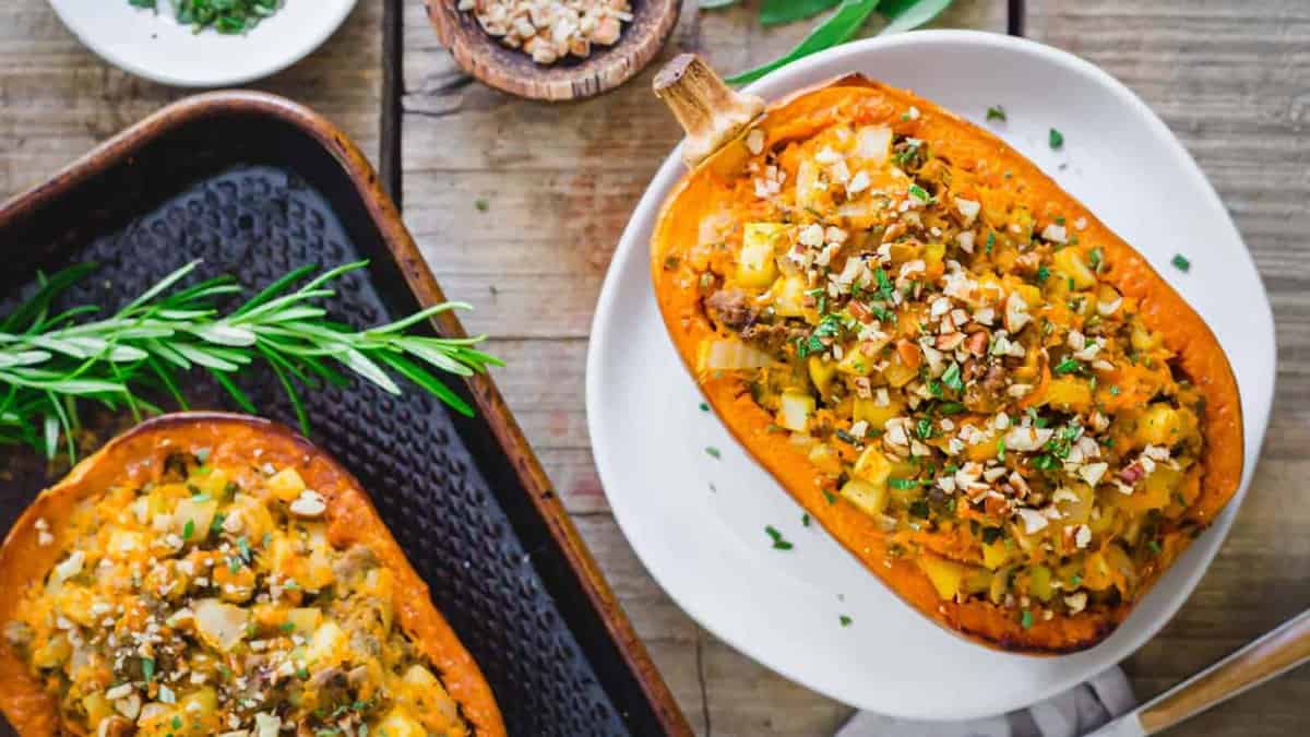 Two halves of stuffed butternut squash filled with vegetables and topped with chopped nuts and herbs are served on plates, with rosemary garnish nearby.