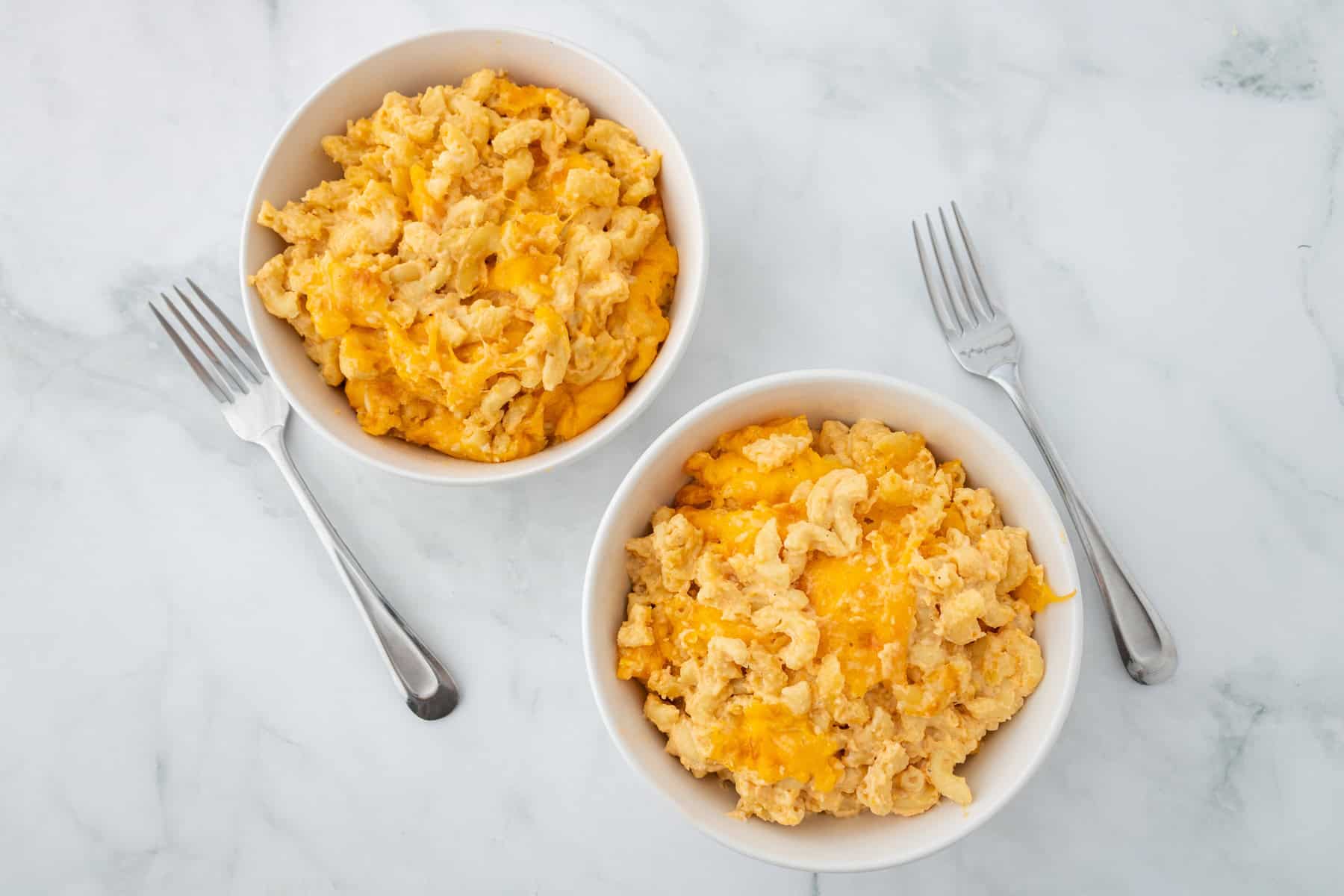 Two white bowls filled with macaroni and cheese sit on a marble surface, each with a metal fork beside them.