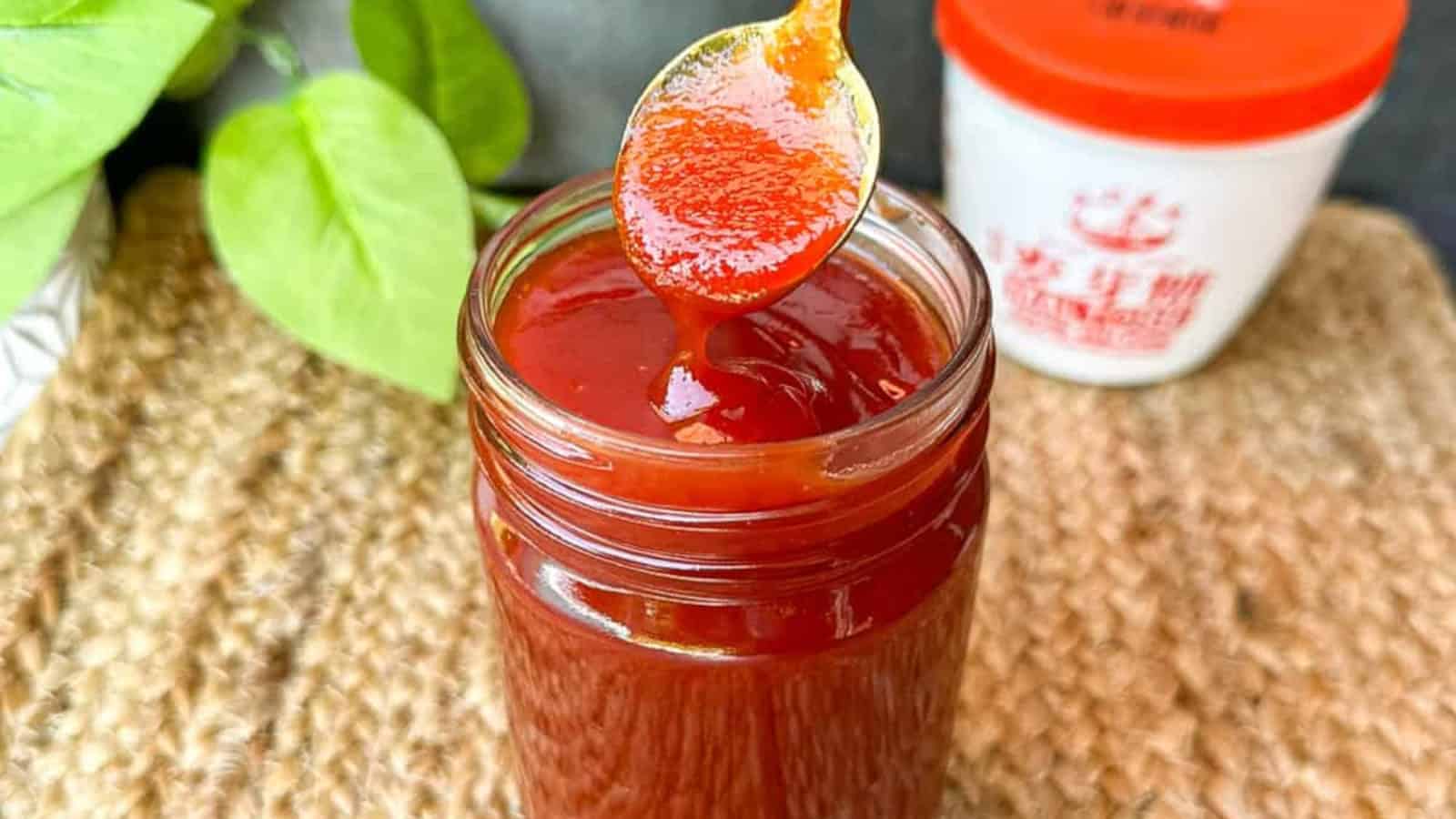 A spoon lifts thick red chili sauce from a glass jar, with a white container and green leaves in the background.