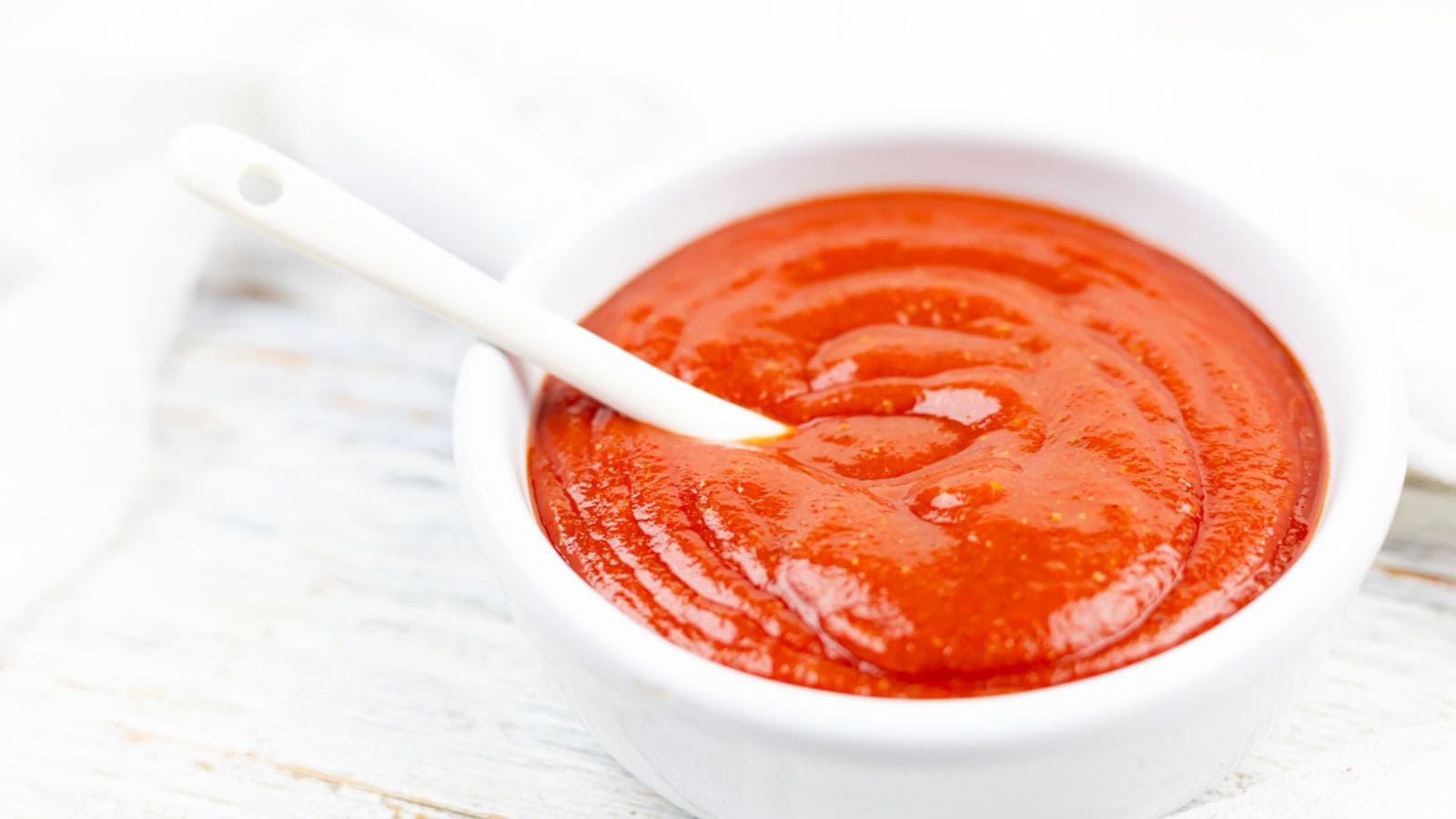 A white bowl filled with smooth red tomato sauce or ketchup, with a white spoon resting inside, on a light wooden surface.