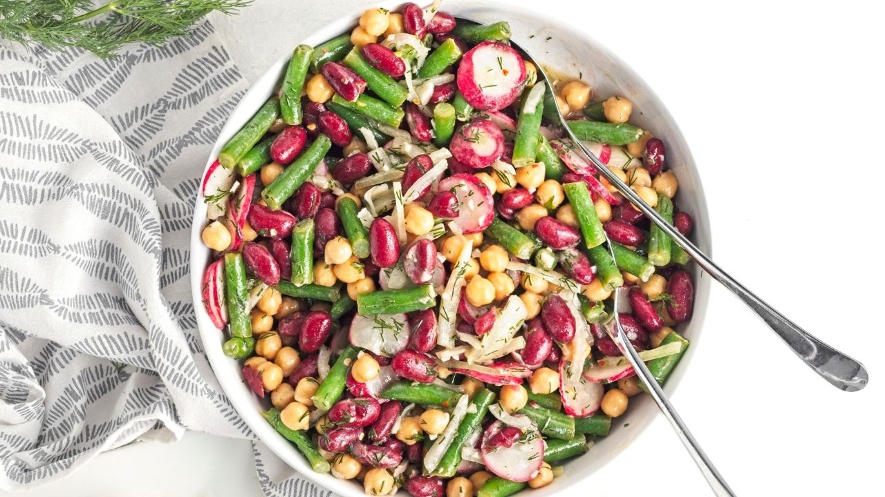 A bowl of mixed bean salad with chickpeas, kidney beans, green beans, radish slices, and onions, with two serving spoons.