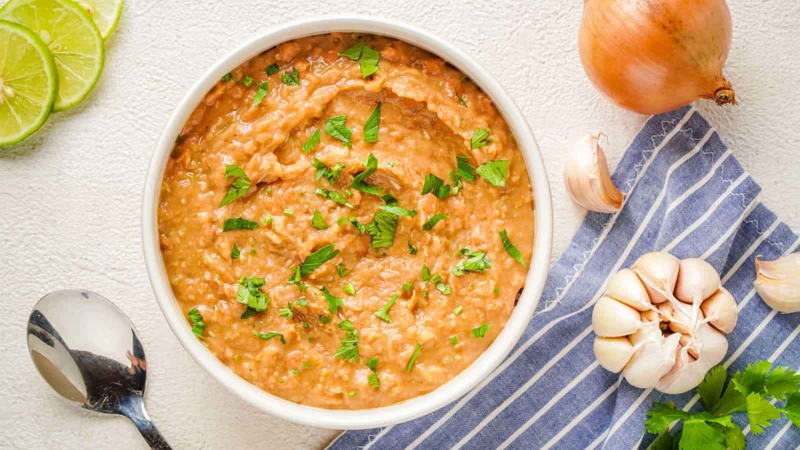 Bowl of refried beans garnished with chopped cilantro, next to a spoon, sliced lime, garlic bulb, onion, and a striped cloth on a white surface.