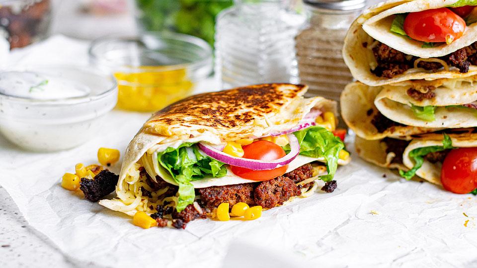 A grilled wrap filled with ground meat, corn, lettuce, tomato, red onion, and cheese, with more wraps and small bowls of condiments in the background.