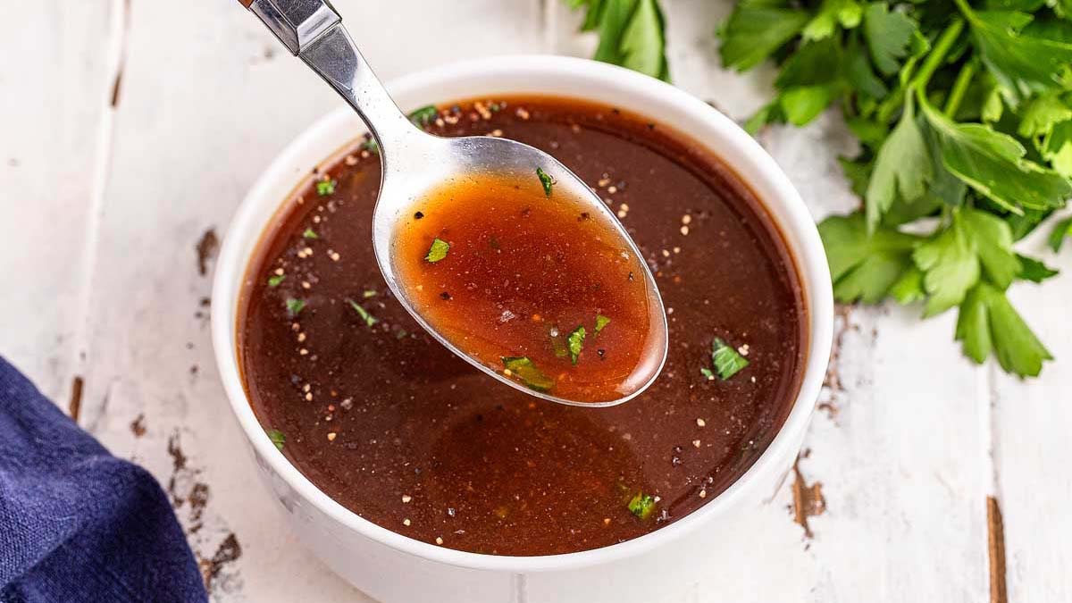 A bowl of brown broth with herbs, with a spoon lifting some broth above the bowl, and parsley in the background.