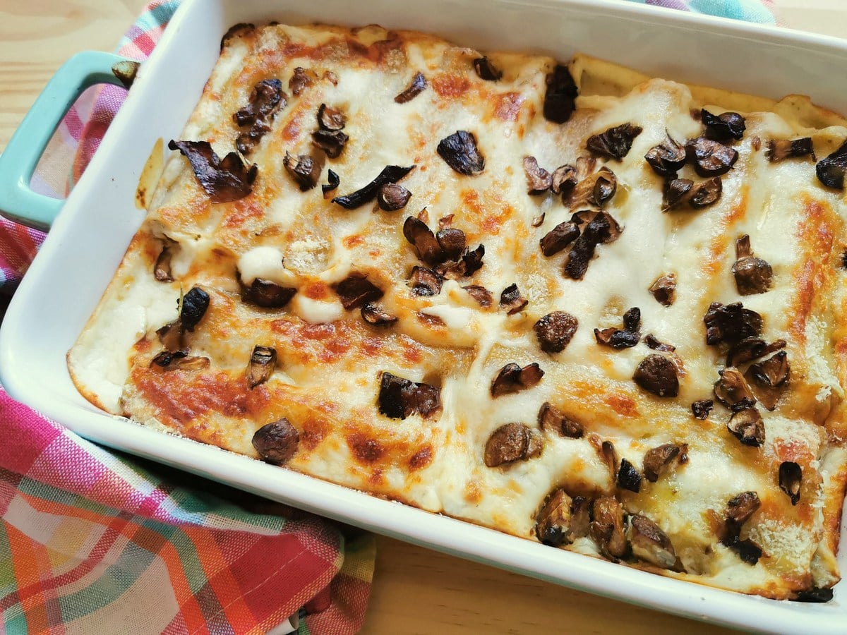 Baked lasagna in a rectangular dish topped with melted cheese and pieces of mushroom, placed on a colorful cloth.