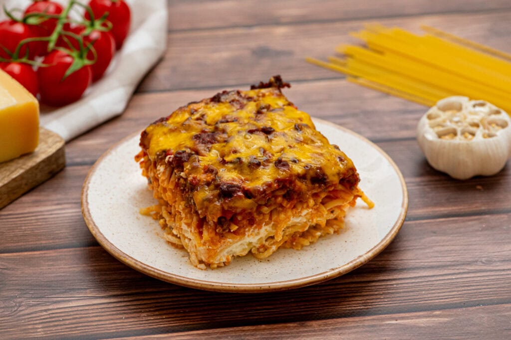 A slice of cheesy baked lasagna on a plate, with raw spaghetti, garlic, cheese, and tomatoes in the background on a wooden table.