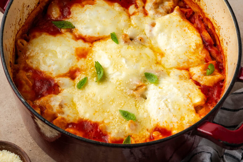 A close-up of baked lasagna in a red pot, topped with melted cheese, tomato sauce, and a few fresh basil leaves.