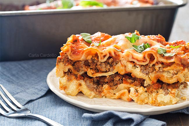 A slice of baked lasagna with layers of pasta, meat sauce, and cheese sits on a white plate, garnished with fresh basil. A metal fork and blue napkin are beside the plate.