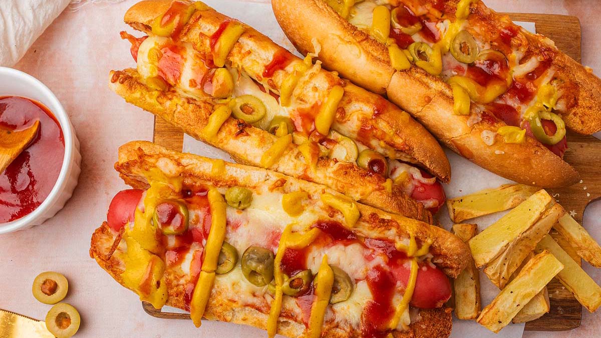 Three hot dogs topped with melted cheese, sliced green olives, ketchup, and mustard are served with a side of fries.