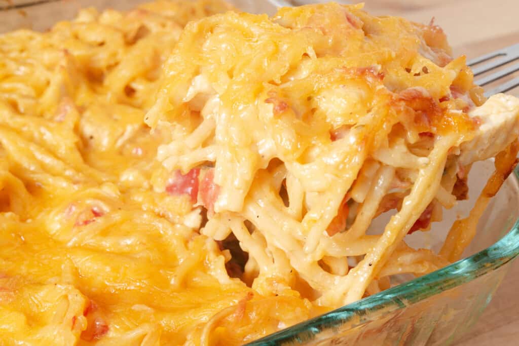 Cheesy baked spaghetti casserole being served from a glass baking dish, with melted cheese and visible pieces of tomato.