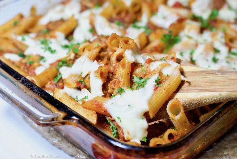 A glass baking dish filled with baked ziti pasta in tomato sauce, topped with melted cheese and chopped parsley, with a wooden spatula lifting a portion.