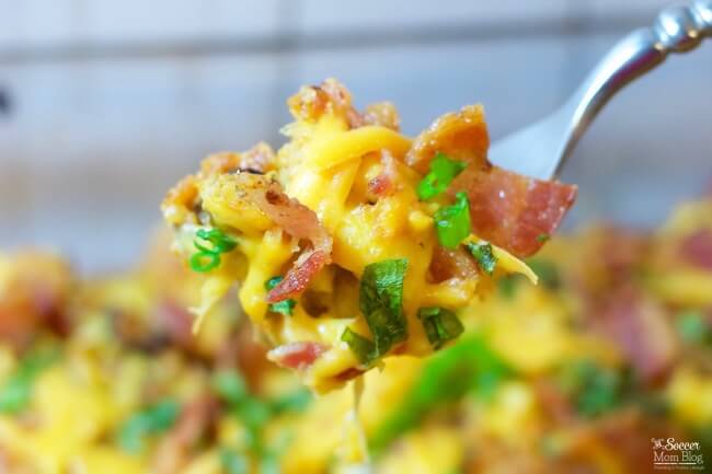 A fork holds a bite of cheesy casserole with pieces of bacon and chopped green onions.