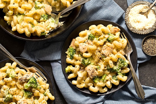 Three plates of creamy pasta with broccoli and chicken are served on a dark table with forks, next to bowls of grated cheese and black pepper.