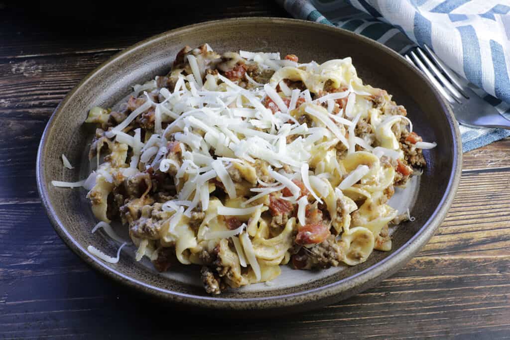 A plate of pasta with ground meat, tomatoes, and shredded cheese on top, set on a dark wooden table with a striped napkin and fork nearby.
