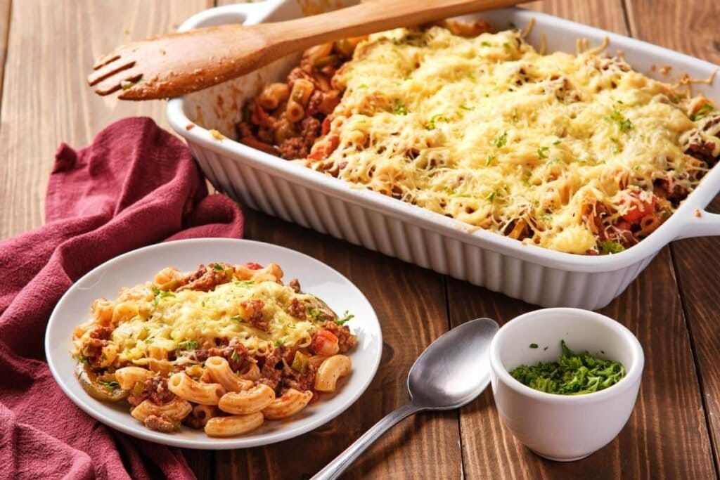 A plate and baking dish of baked pasta with ground meat, tomato sauce, and melted cheese sits on a wooden table beside a spoon, small bowl of herbs, and a red napkin.