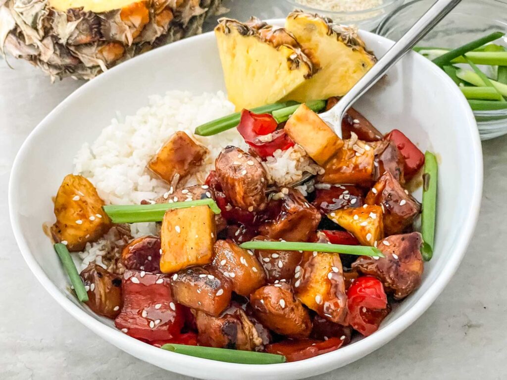 A bowl of white rice topped with teriyaki chicken, pineapple chunks, red bell peppers, green onions, and sesame seeds, with pineapple slices in the background.