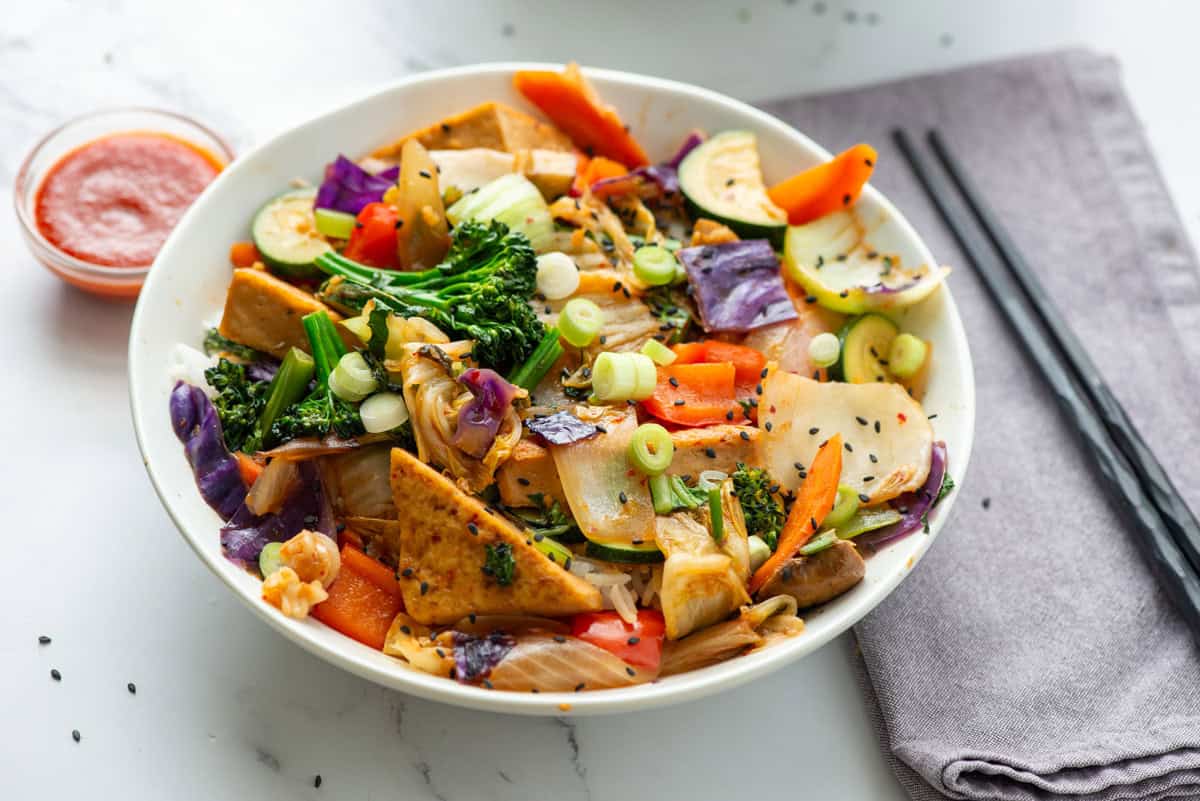 A bowl of colorful stir-fried vegetables and tofu garnished with green onions and black sesame seeds, with chopsticks and a small dish of sauce on the side.