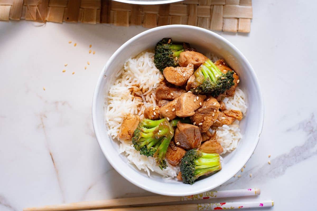 A bowl of white rice topped with cooked chicken pieces, broccoli florets, and sesame seeds. Wooden chopsticks rest beside the bowl on a marble surface.